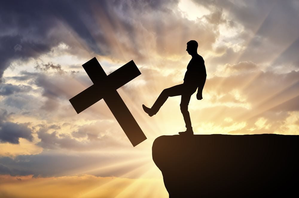 Man atheist pushes the symbol of christian cross into the cliff on sunset background. Concept of atheism