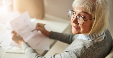 Portrait of a female pensioner in round glasses holding a pen and a document in her hands